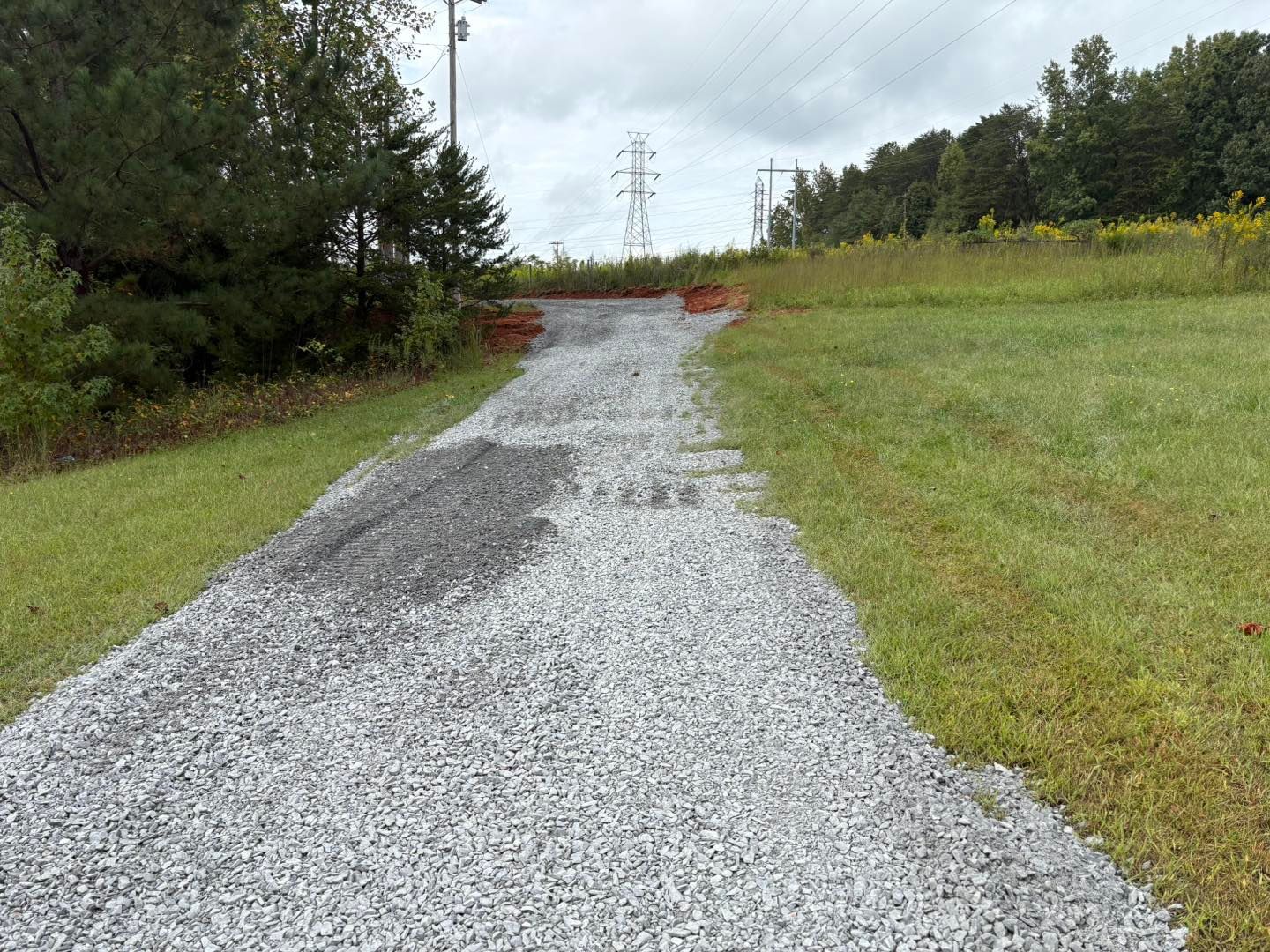 Gravel path sloping upward through grass, leading to trees and power lines on a cloudy day.
