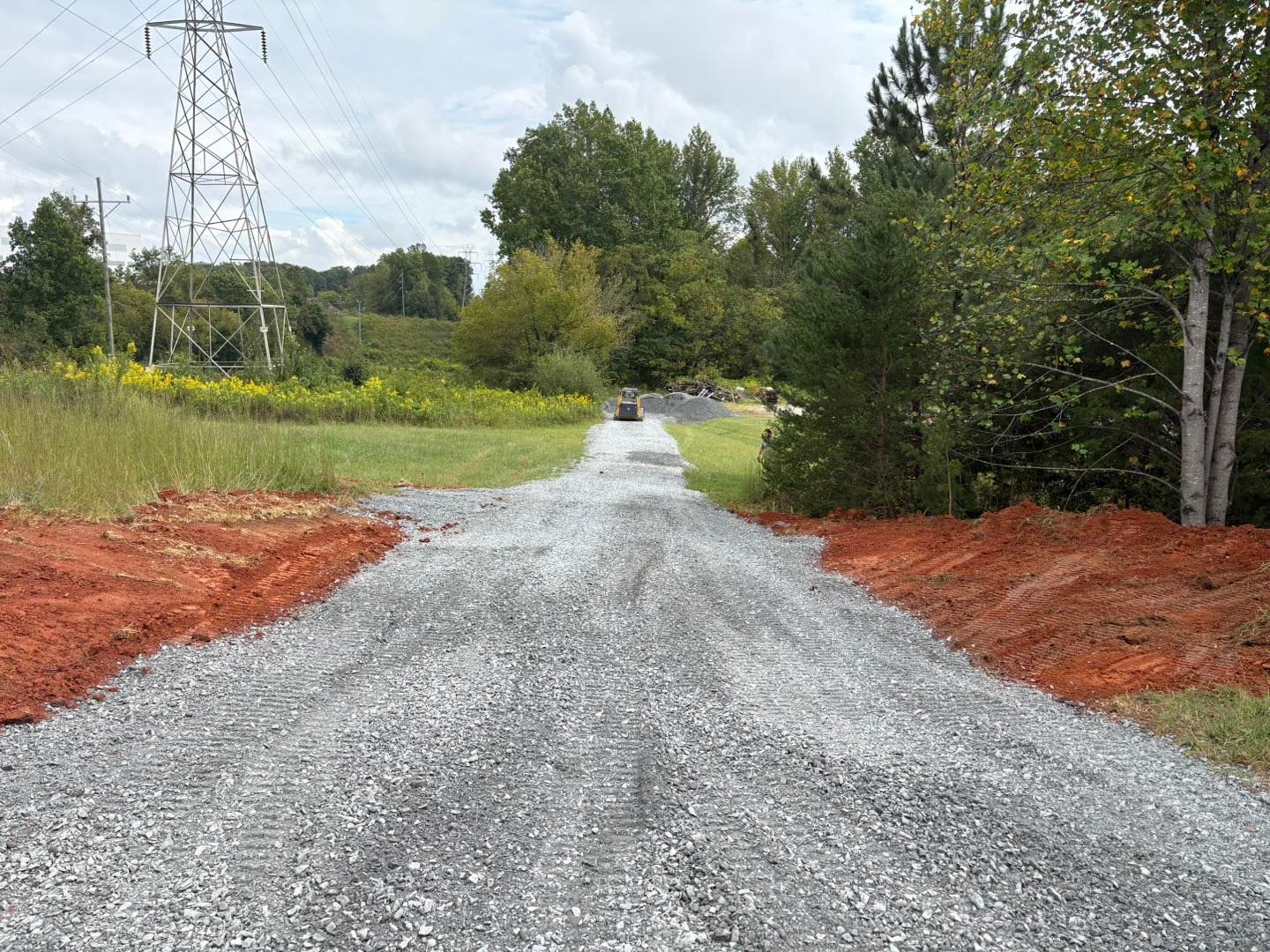 Gravel road with red dirt borders leading toward a green treeline, under a cloudy sky. A power line tower is in the background.