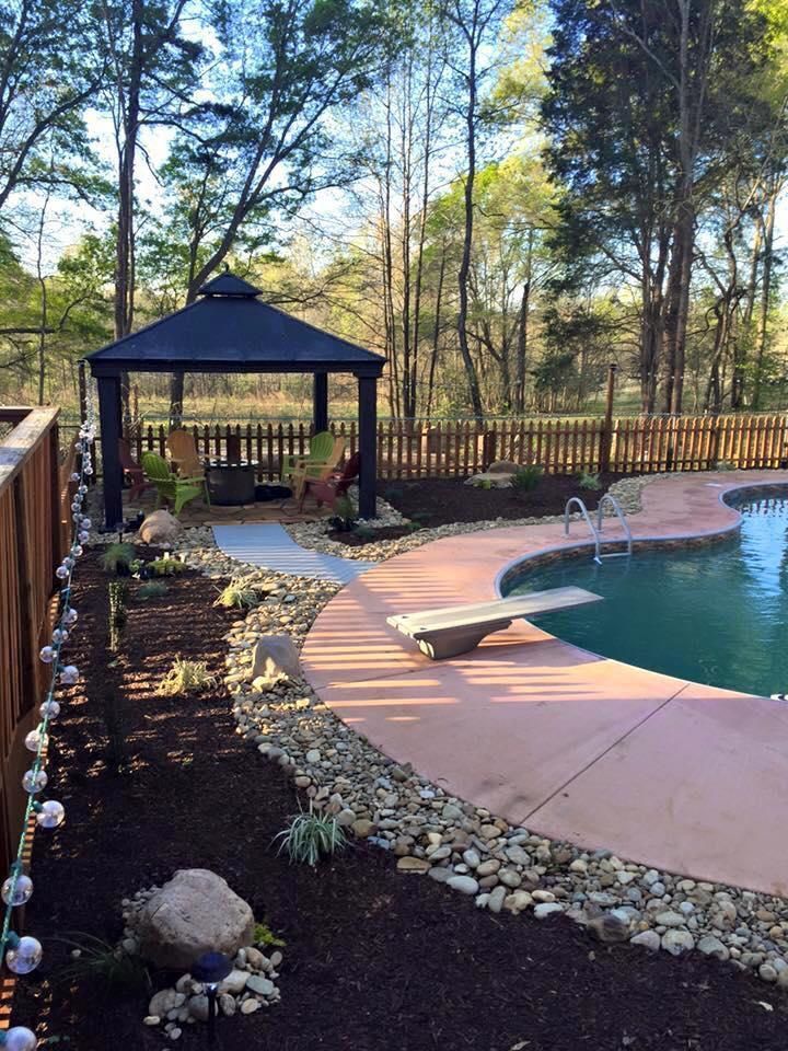 Backyard with pool, gazebo, and stone pathway. Brown mulch and trees surround the area, creating a serene environment.