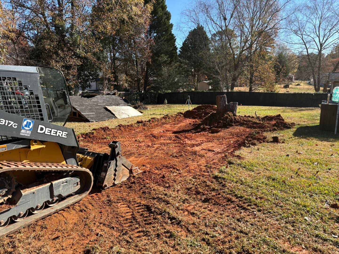 A John Deere skid steer excavates red dirt in a grassy yard, likely for a construction project.