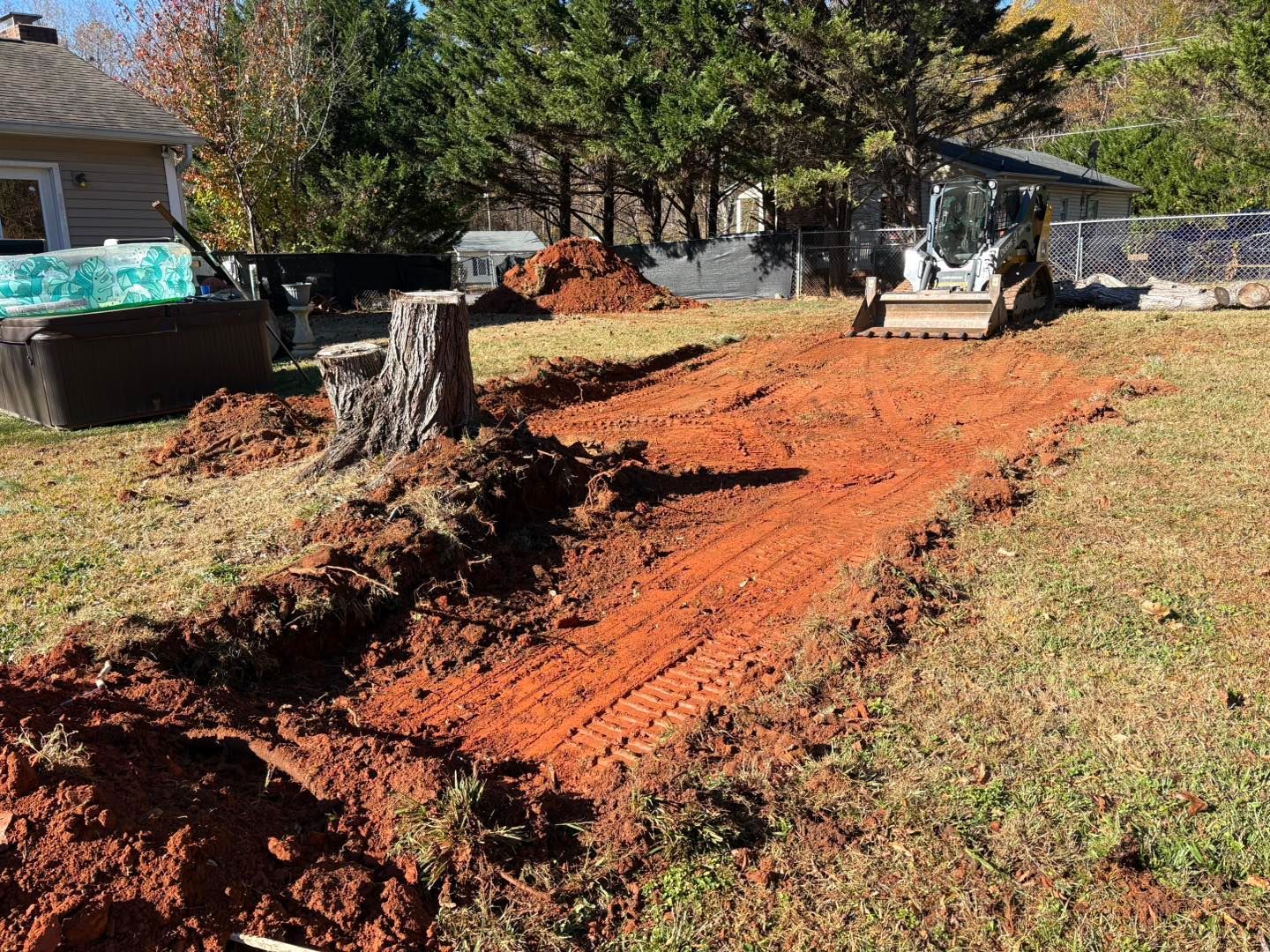 A small bulldozer clearing red soil in a backyard, beside a tree stump.