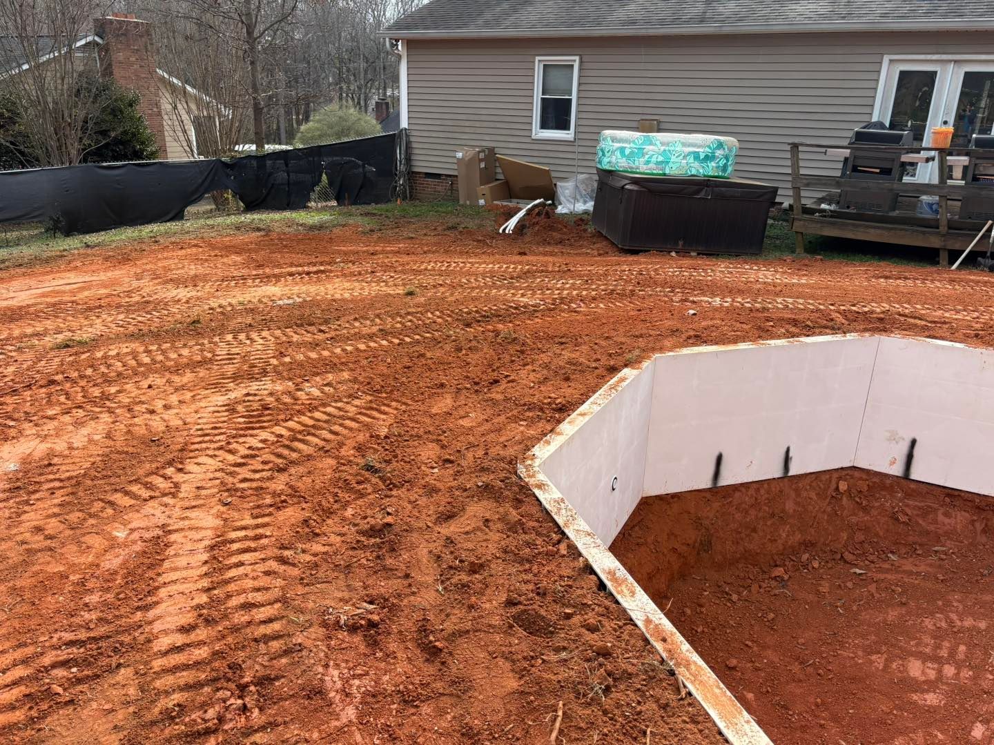 A construction site with a red dirt ground. A white, octagonal structure is in the foreground. A house is in the background.
