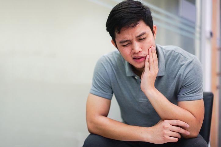 Man in a gray shirt clutching his cheek in pain while sitting indoors