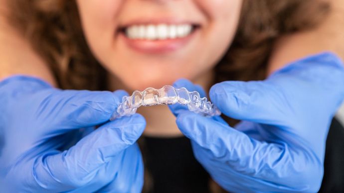 Blue-gloved hands holding a clear dental aligner in front of a smiling mouth