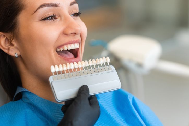 Woman smiling while holding a shade guide for teeth whitening in a dental setting