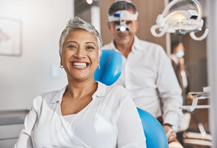 Smiling dental patient in white blouse, seated in clinic chair with dentist and blue exam light behind her