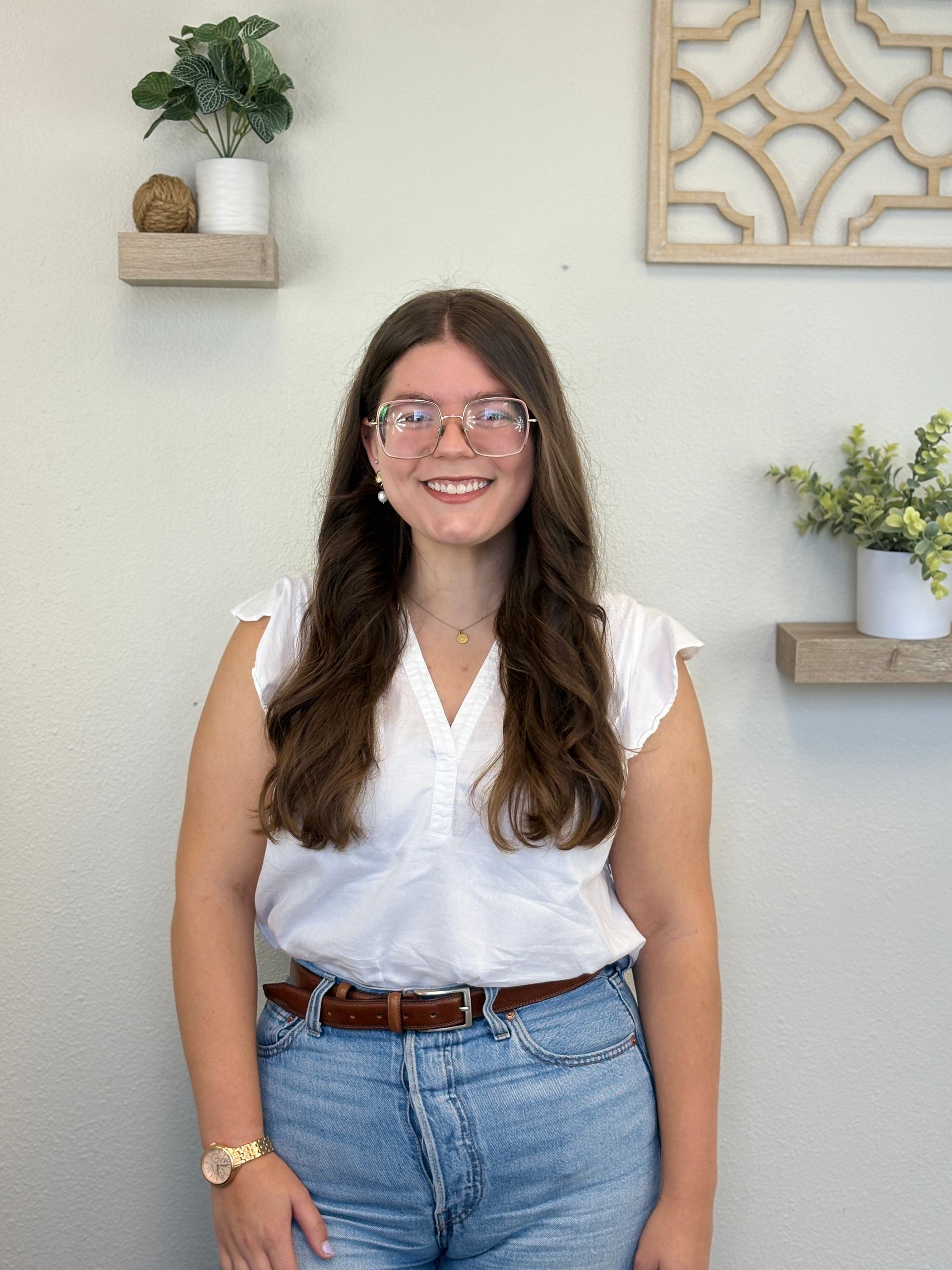 Woman wearing glasses, white top, jeans, and a brown belt smiles in a room with shelves and plants.