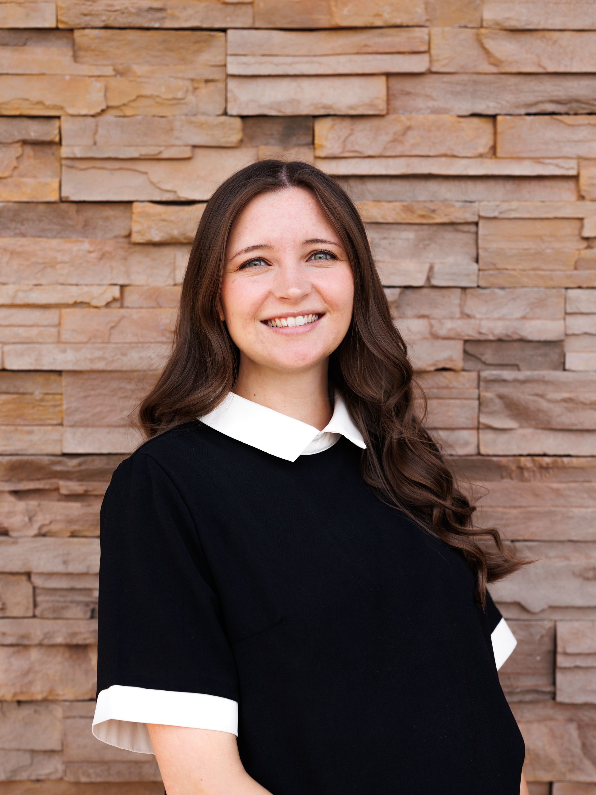 Woman with long brown hair smiles at the camera, wearing a black dress with a white collar, against a stone wall.
