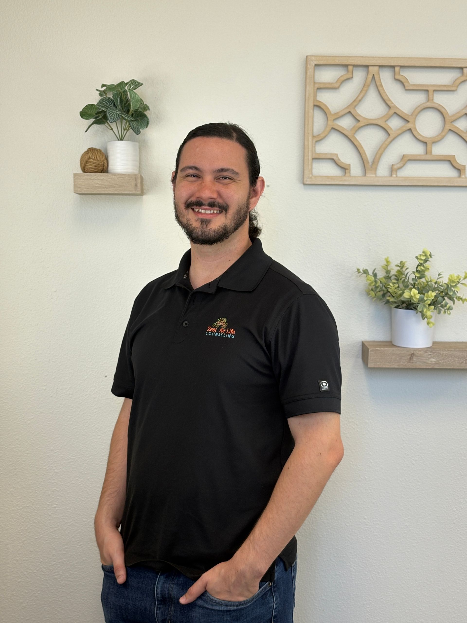 Man with beard and smile in black polo shirt, hands in pockets, standing in front of white wall with decor.