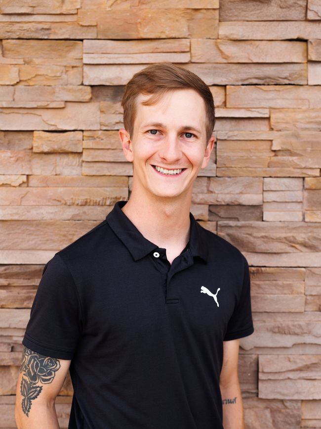 Man in black polo shirt smiling, with arm tattoo, standing in front of brick wall.