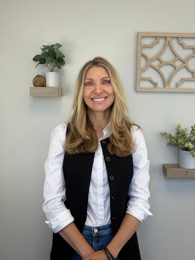 Woman in a black vest smiles; standing in front of a wall with shelves, plants, and decorative frame.