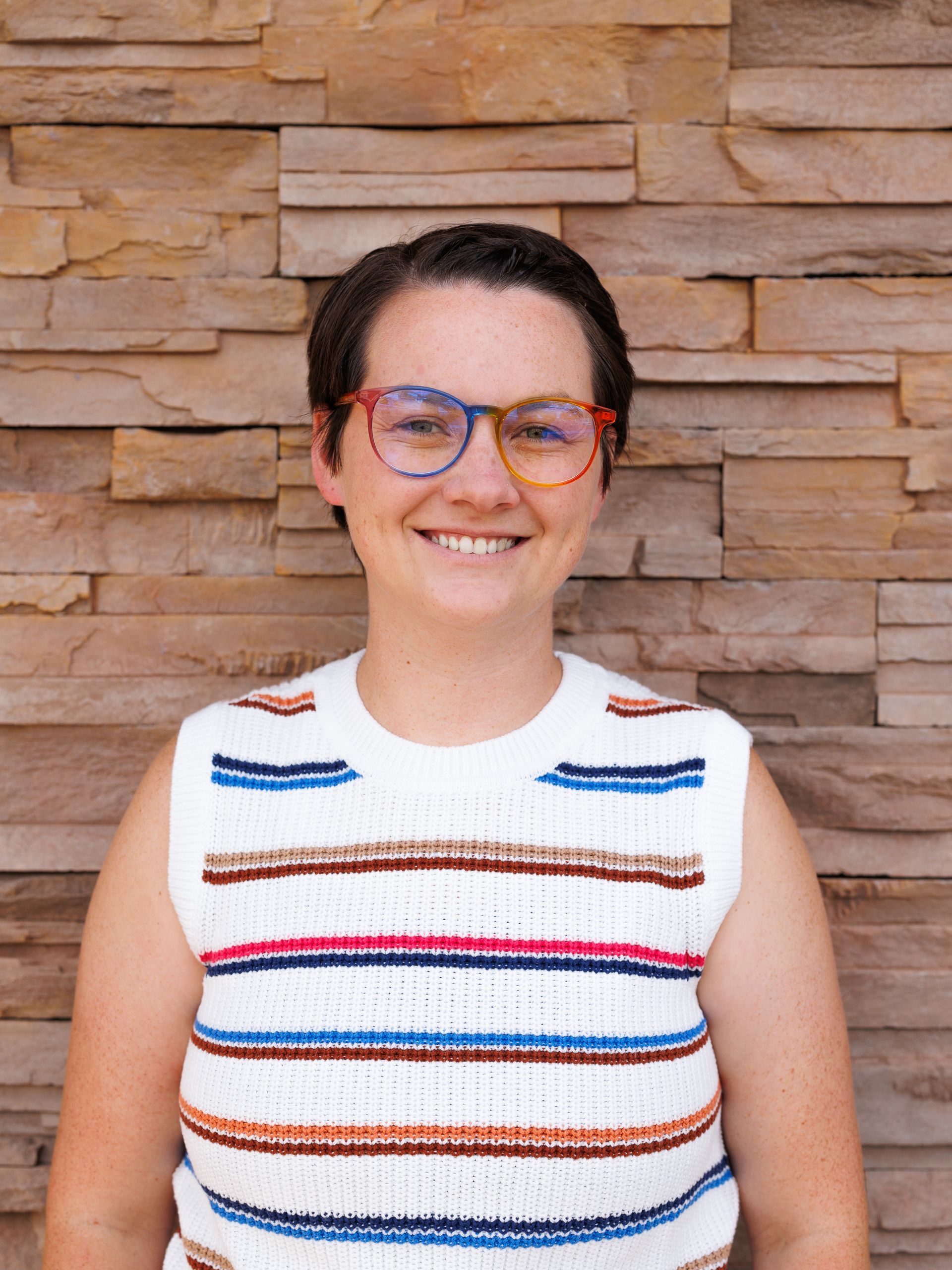 Woman with short dark hair and glasses, wearing a striped vest, smiling in front of a brick wall.