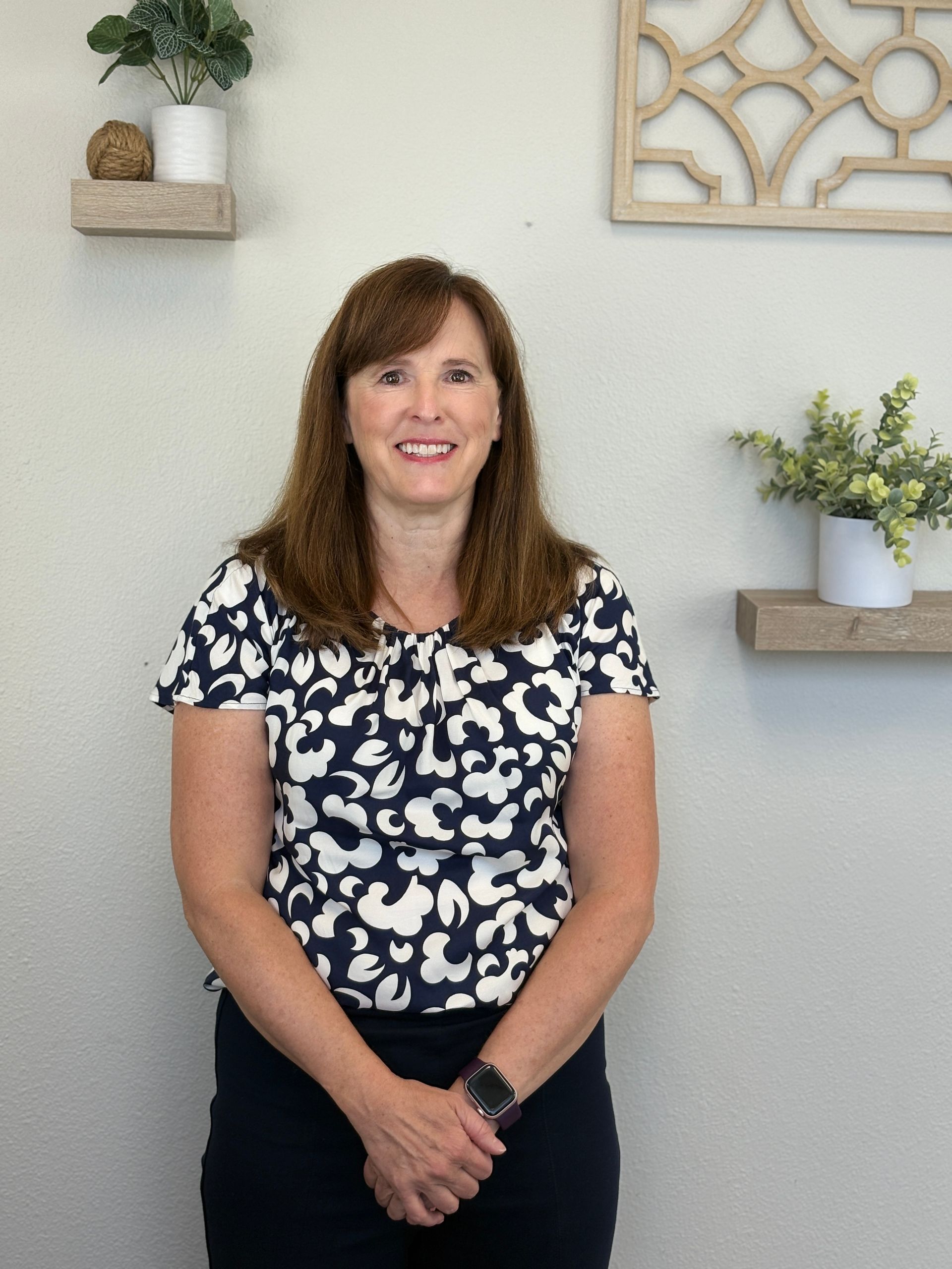 Woman with brown hair in a navy and white top, standing in front of a wall with shelves holding plants.