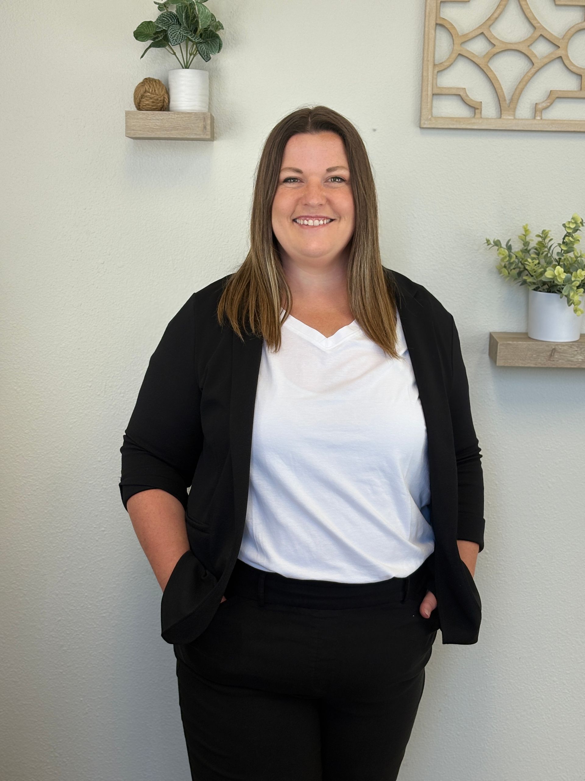 Woman smiling, wearing a black blazer and pants over a white shirt; standing against a wall with plants.
