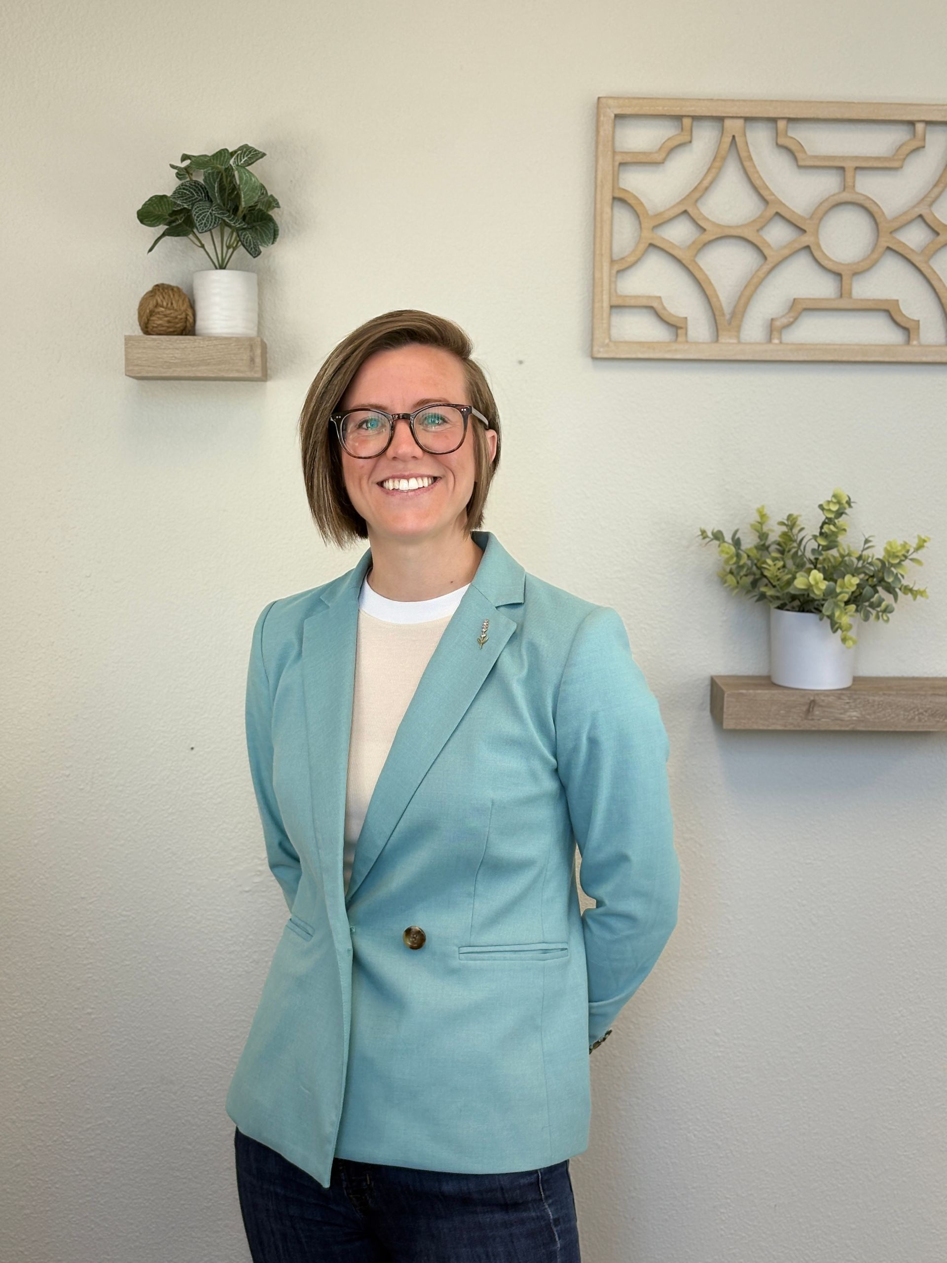 Woman in teal blazer, glasses, smiles, hands behind back, standing near wall with plants and decorative wood.