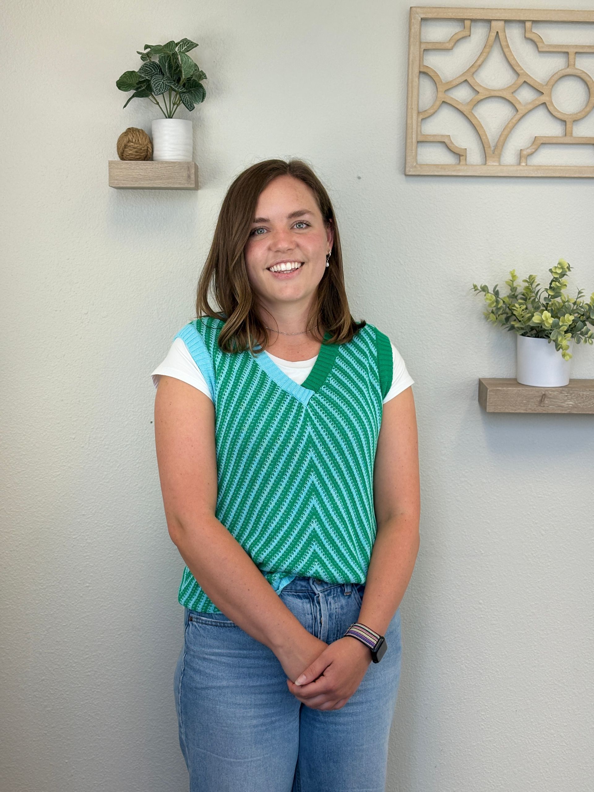 Woman in green chevron vest and jeans smiles in front of shelves with plants.