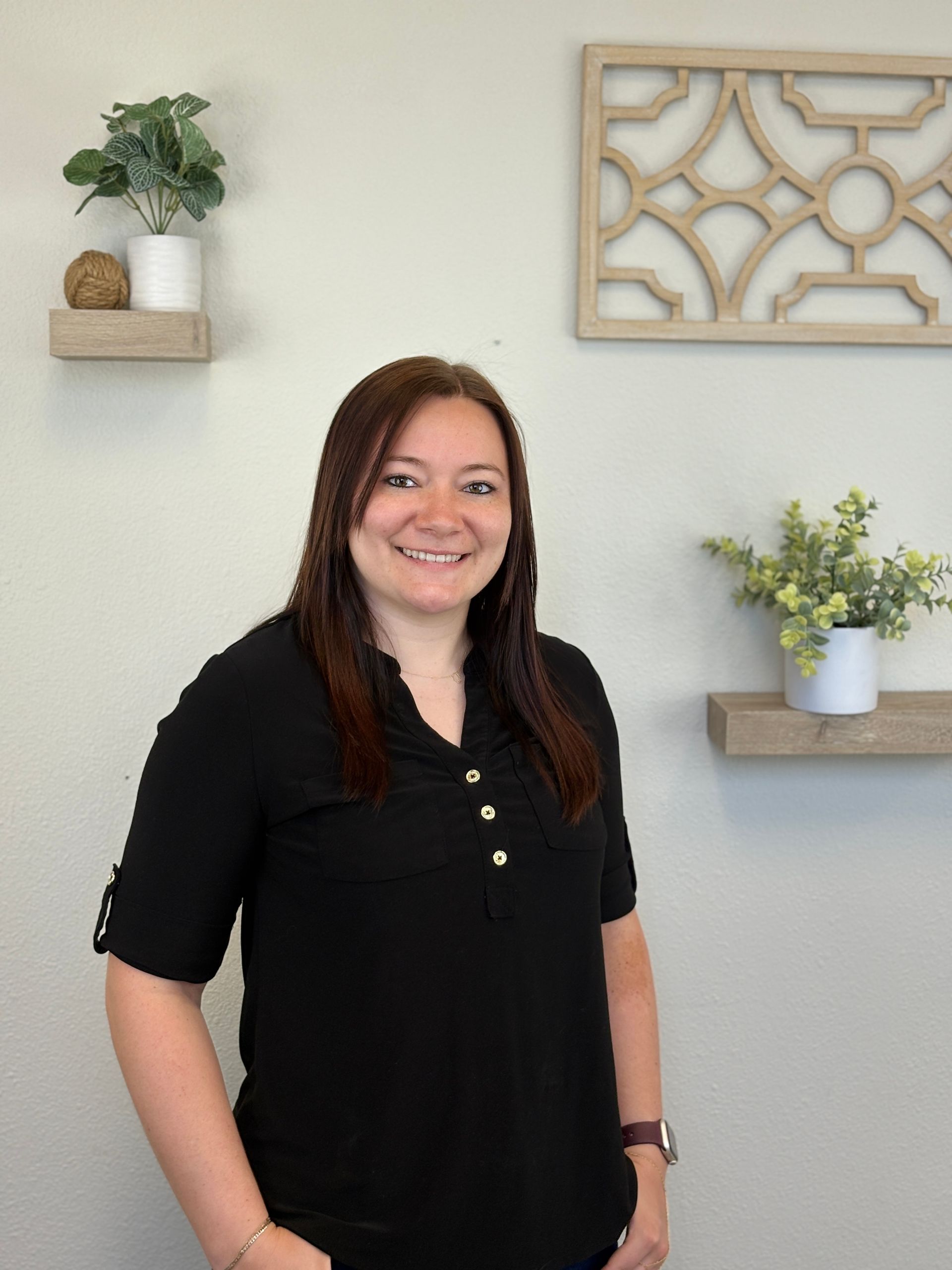 Woman with brown hair smiles, standing in front of a light wall decorated with shelves and plants.