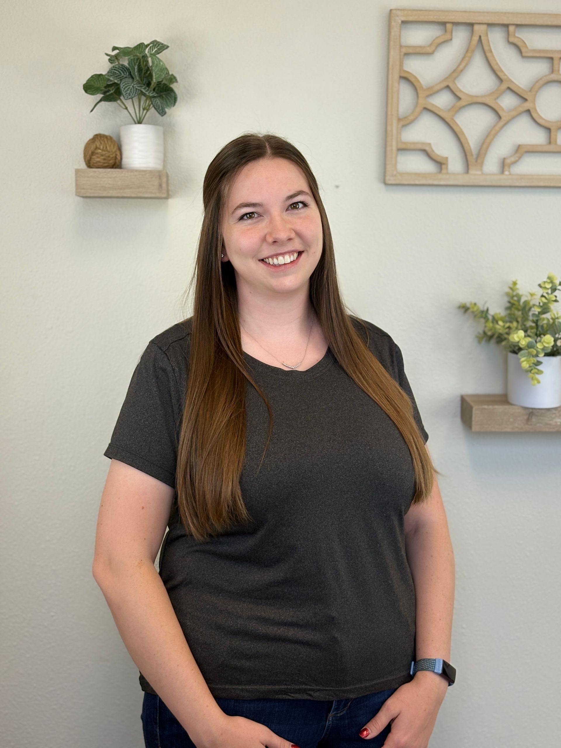 Woman with long brown hair smiling in front of shelves with plants, wearing a gray t-shirt.