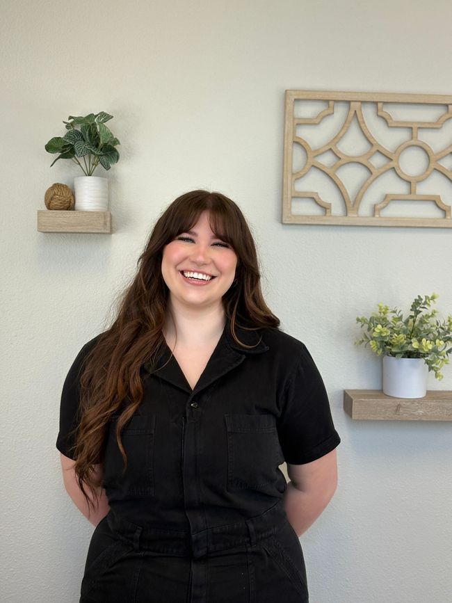 Woman with long brown hair smiles in front of a wall with shelves holding plants and a decorative wooden piece.