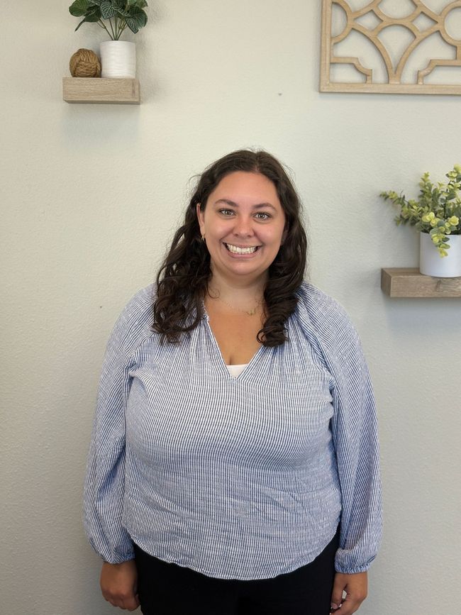 Woman with curly hair, smiling, wearing a blue and white striped blouse, standing in front of a white wall with shelves.