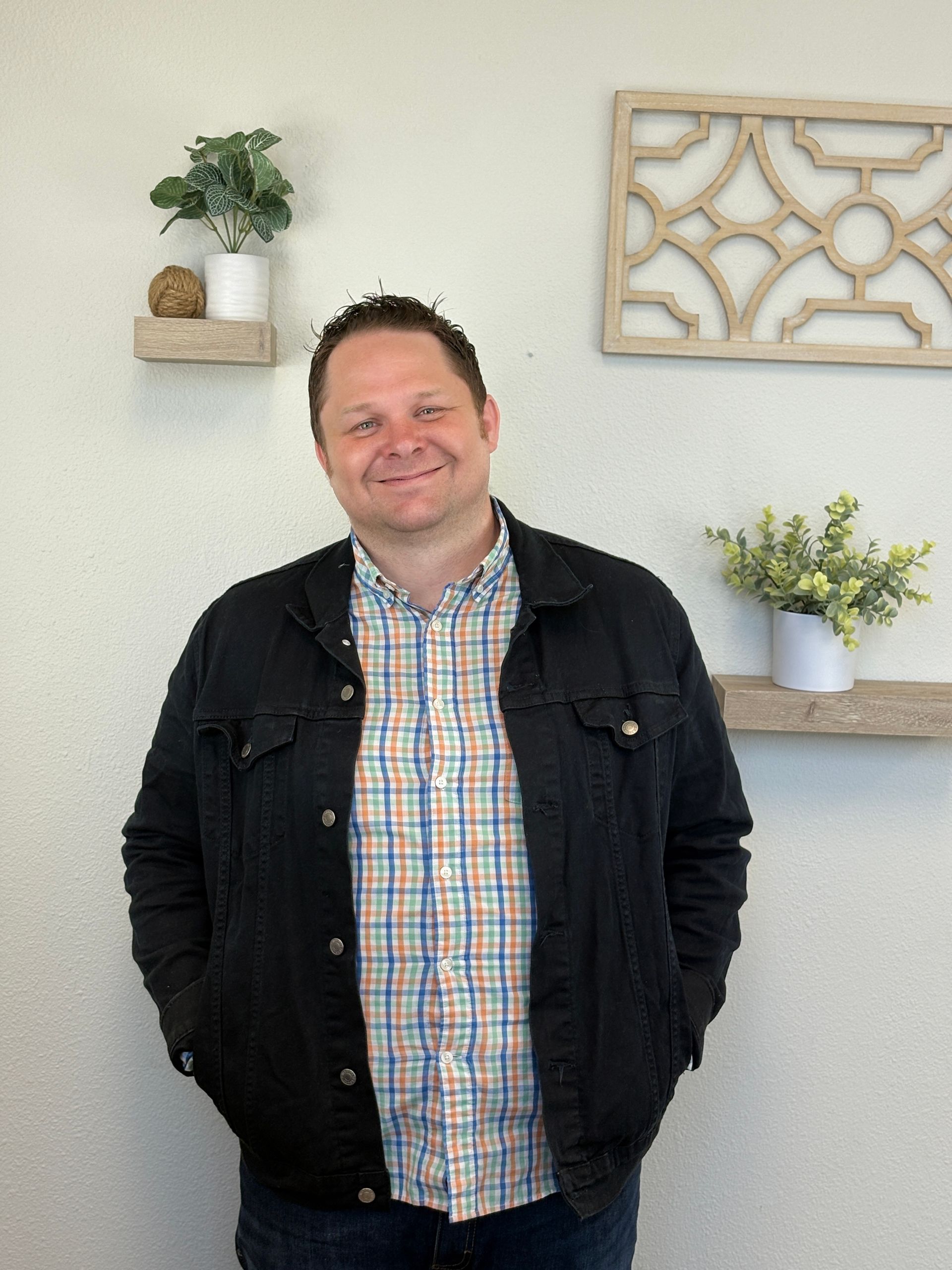 Man in denim jacket smiles, stands indoors, hands in pockets.