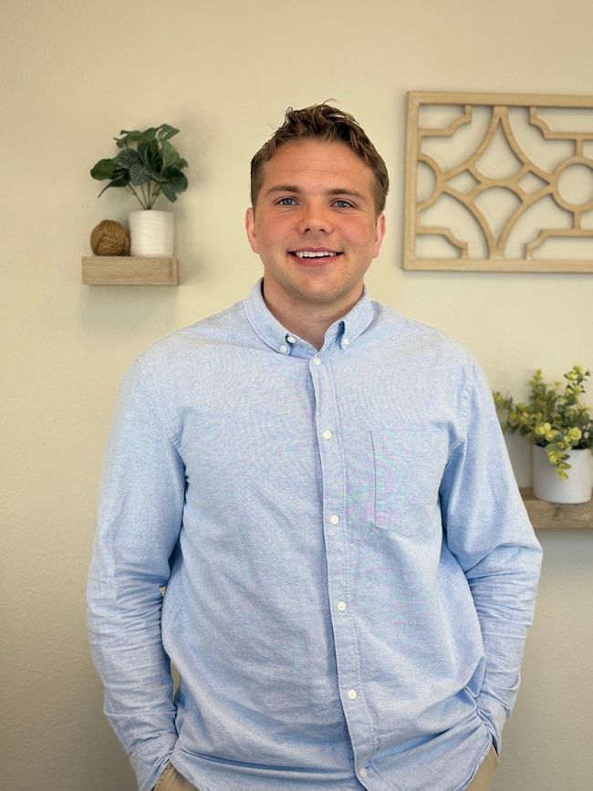 Man in blue button-down shirt smiling, standing indoors. Light-colored wall with decor in the background.