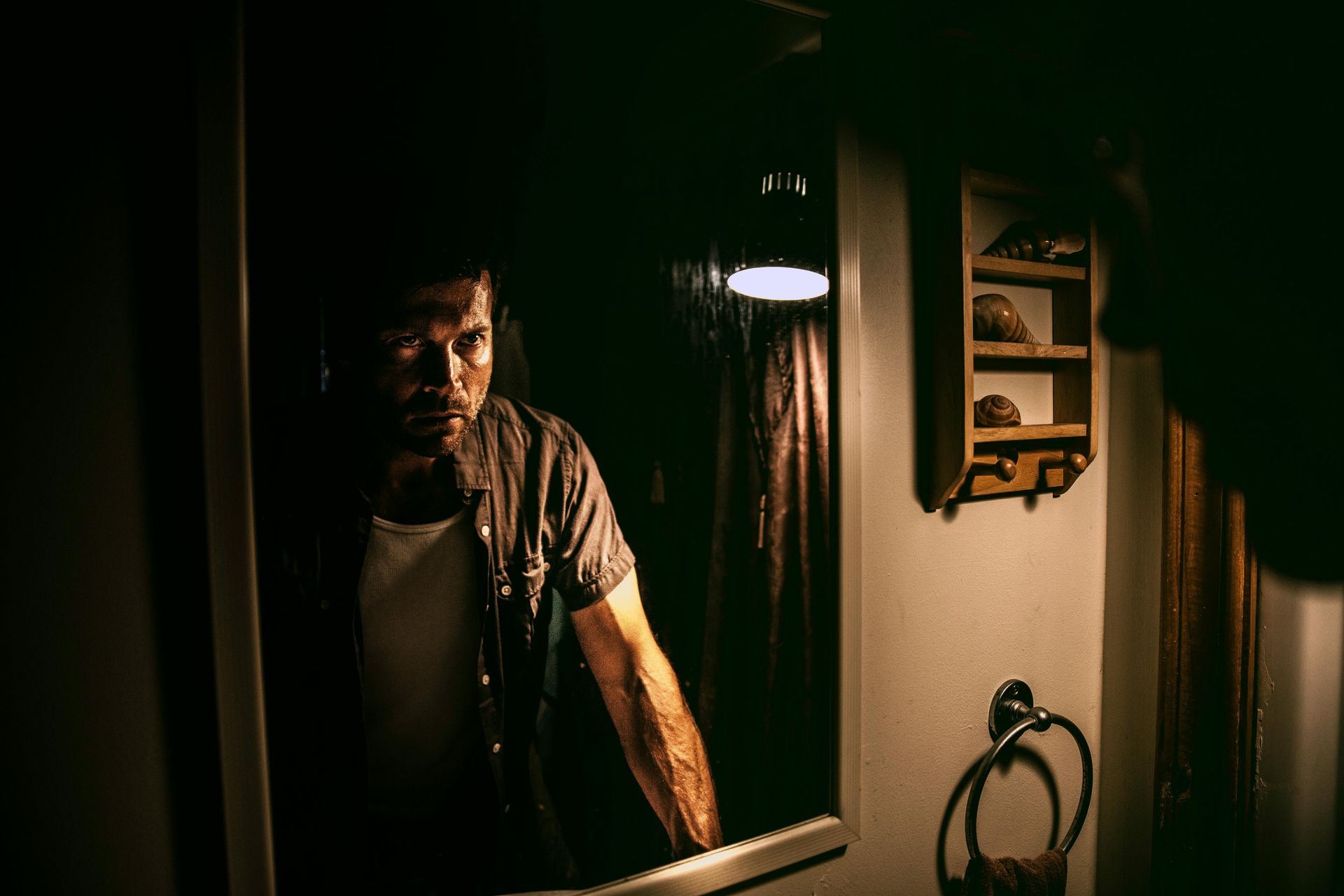 Man looks at himself in a mirror in a dimly lit bathroom.
