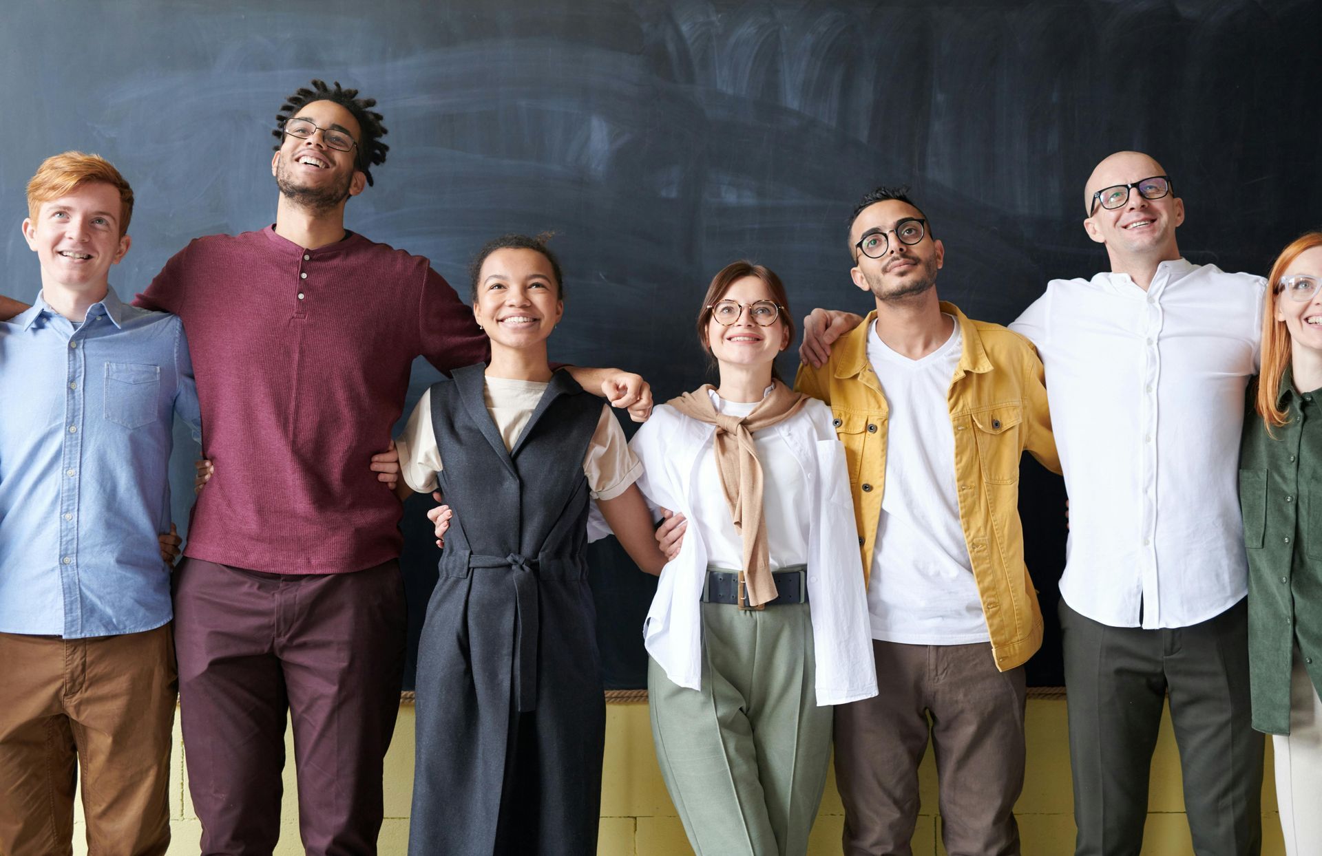 Group of diverse people with arms around each other, smiling, standing in front of a chalkboard.