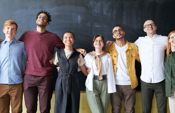 Group of diverse people with arms around each other, smiling, standing in front of a chalkboard.