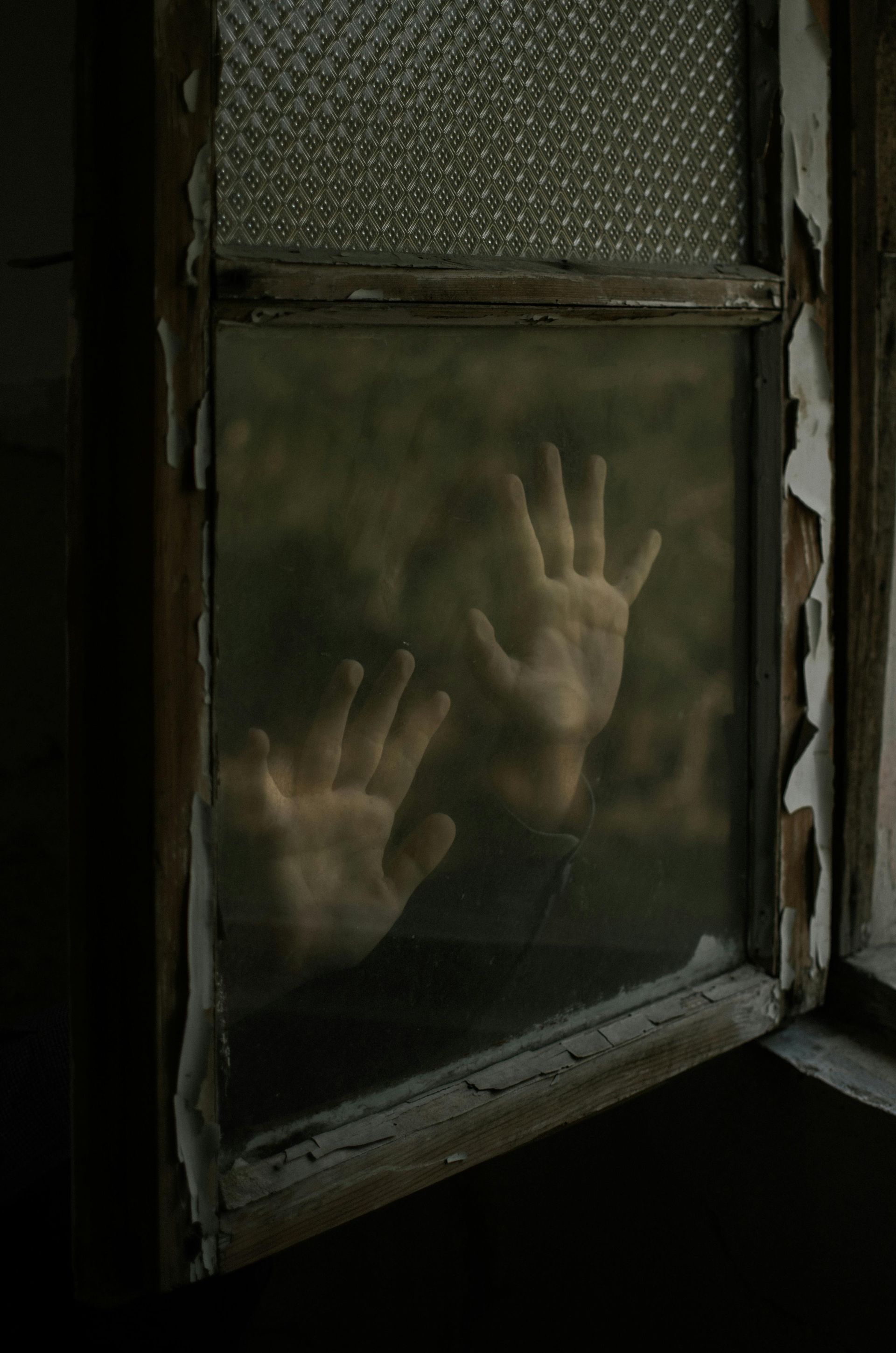Hands pressed against a dirty window, obscured figure. Old wooden frame, peeling paint, dark interior.