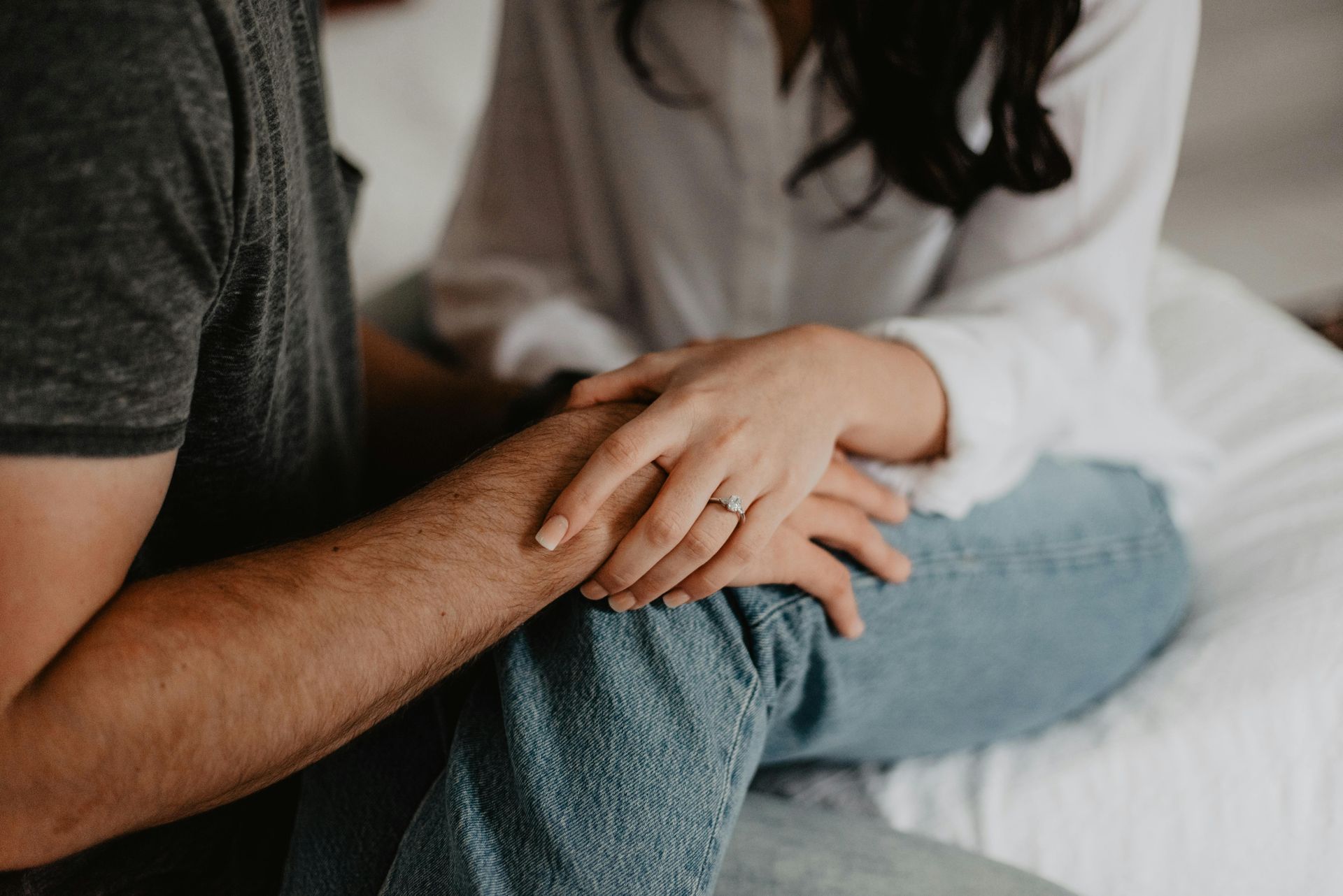 Couple holding hands, woman wearing jeans and a white shirt, engagement ring visible, indoor setting.