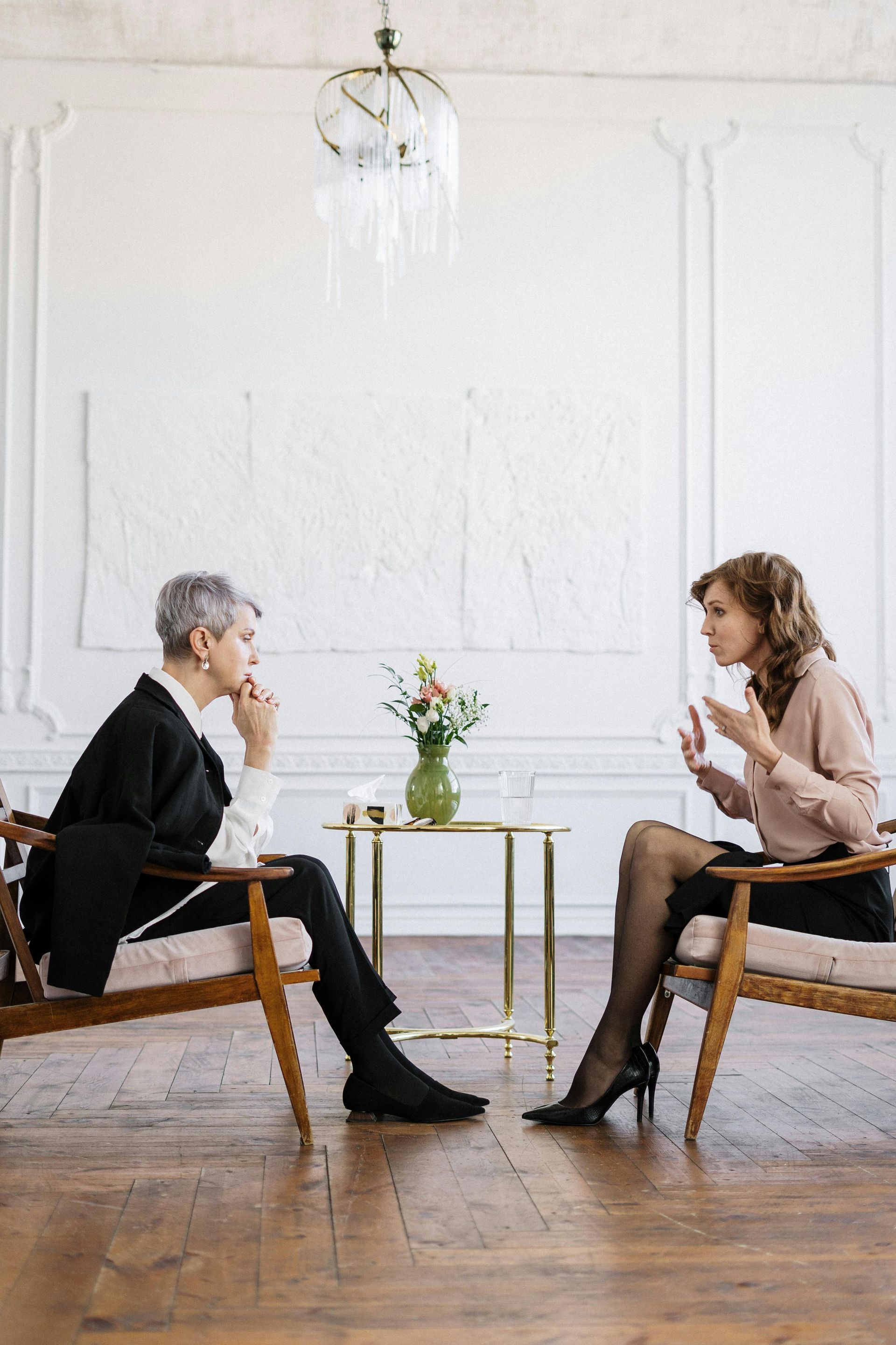 Two people in chairs, talking in a bright room; one gesturing while the other listens thoughtfully.