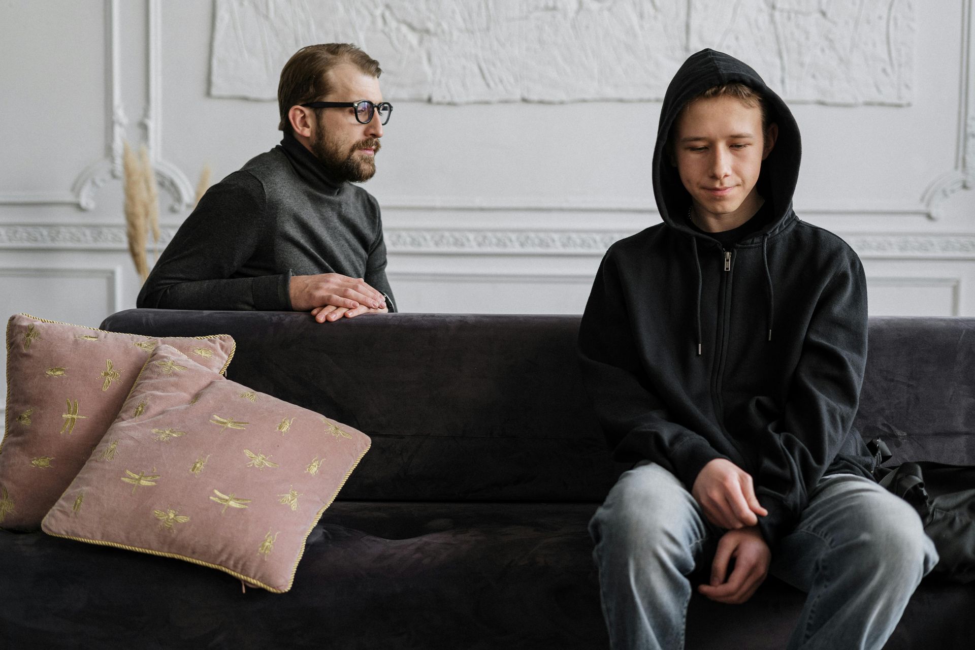 Man with glasses, looking at a teenager wearing a black hoodie, sitting on a couch, looking down.