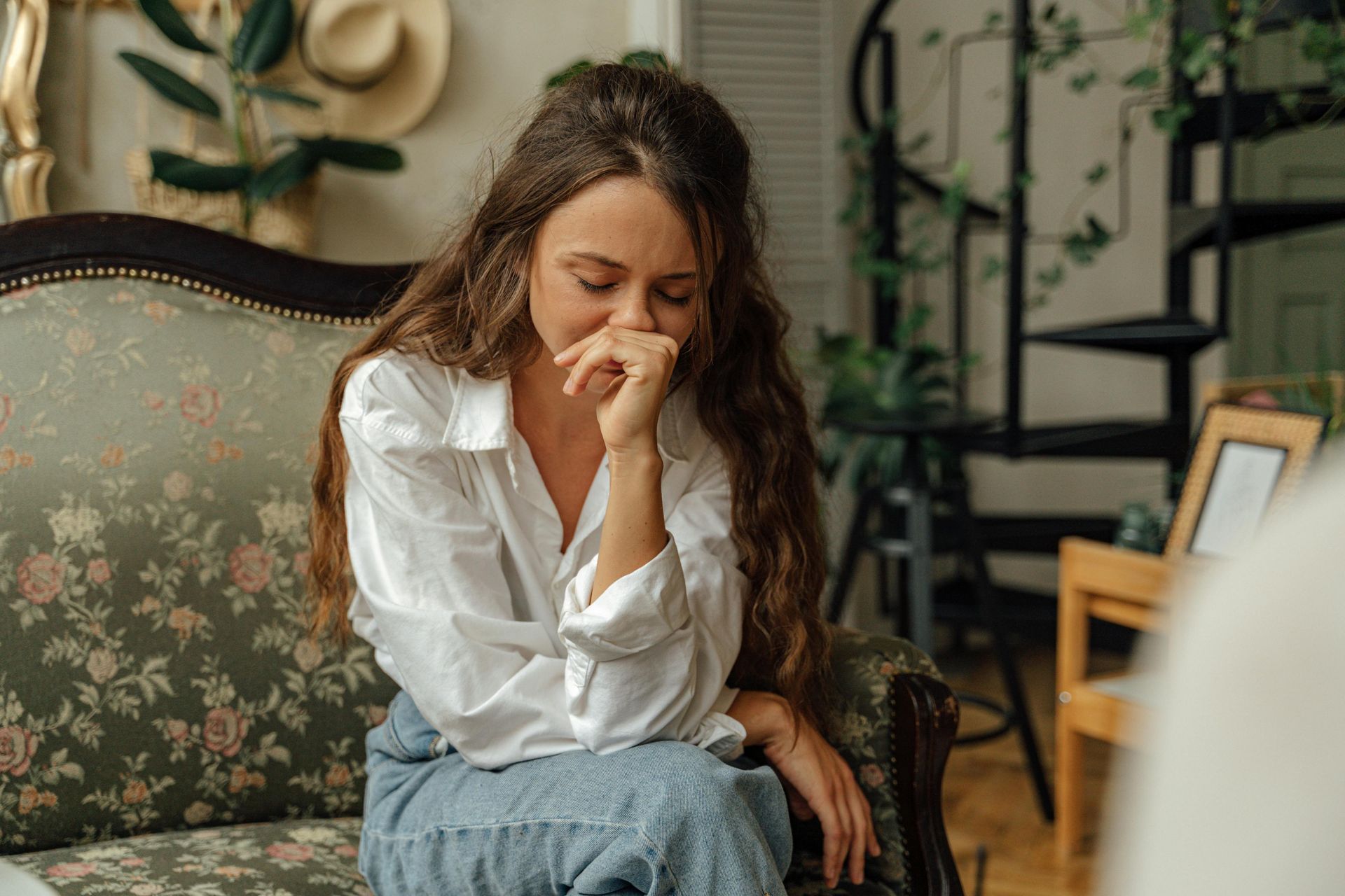 Woman with long brown hair, sitting on ornate sofa, covering mouth with hand; possibly sad.