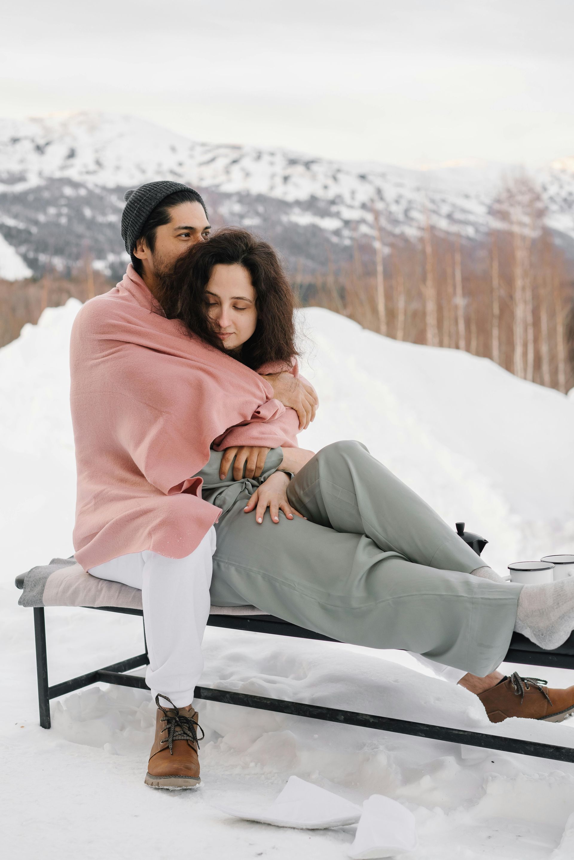 Couple embraces on a snow-covered bench, wrapped in a pink blanket with a mountain backdrop.