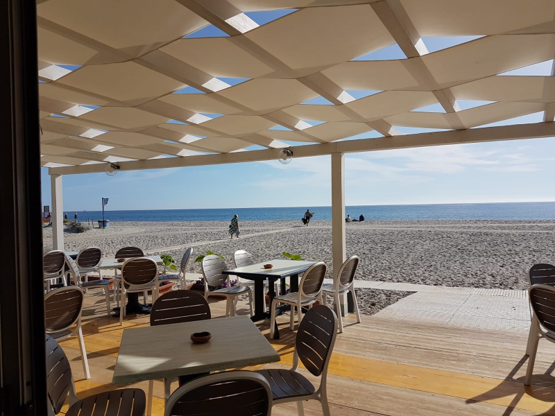 Bar all'aperto con vista su una spiaggia di ciottoli e sull'oceano, sotto una tettoia bianca, in una giornata di sole.