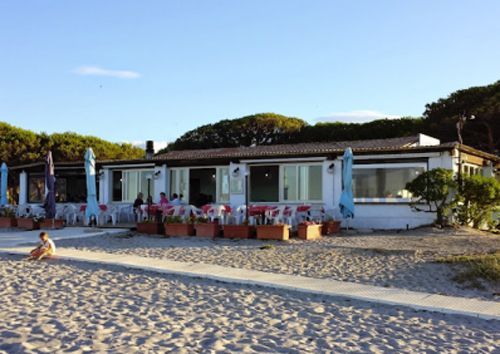 Ristorante sulla spiaggia con posti a sedere all'aperto, edificio bianco, ombrelloni blu, sabbia e alberi.