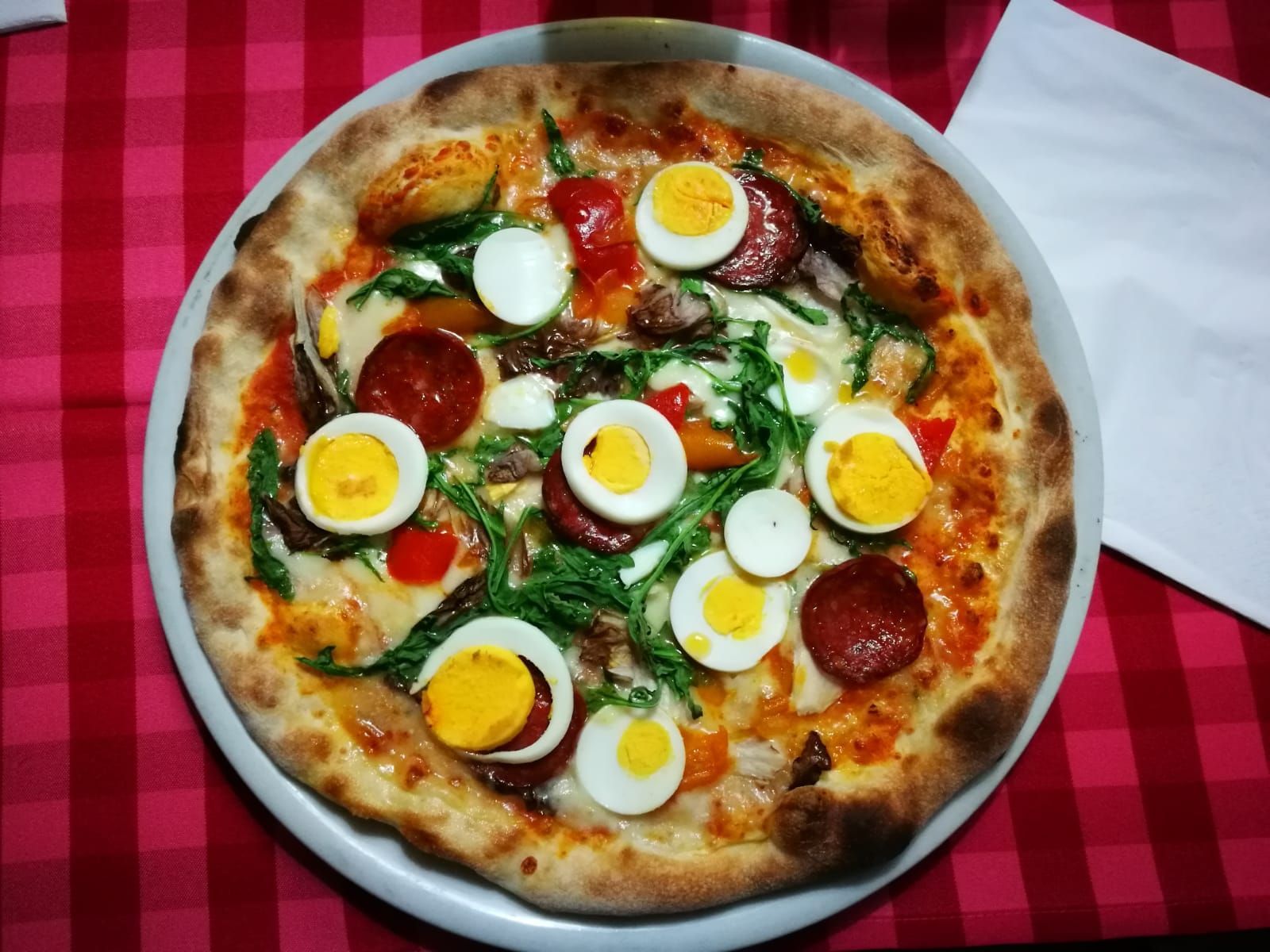 Pizza con uova sode a fette, salame piccante, verdure e peperoni rossi su un piatto bianco, sopra una tovaglia a quadri rossi e bianchi.