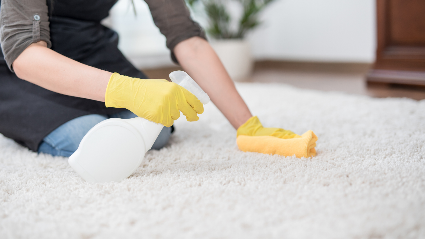 Person wearing yellow gloves sprays and wipes a white carpet with a sponge.