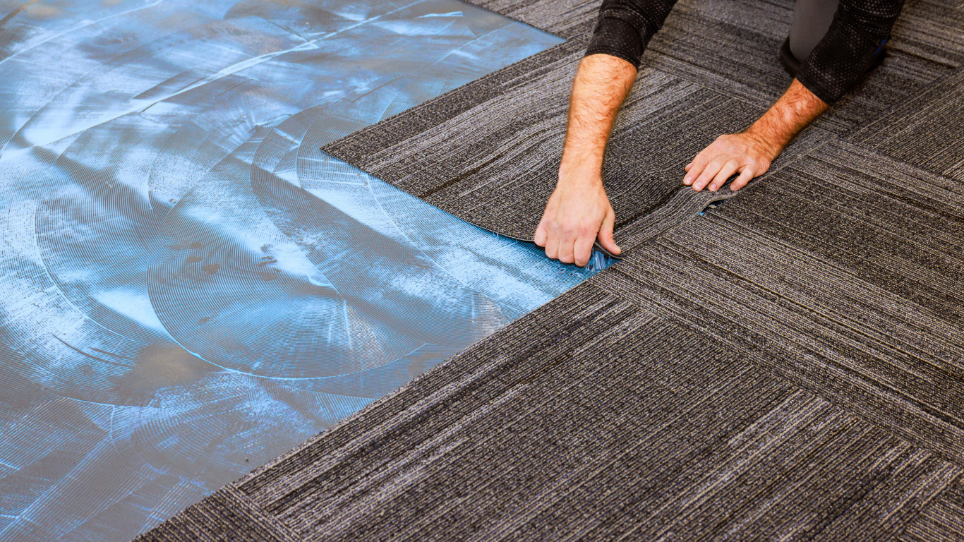 Person installing gray carpet tiles over a blue adhesive layer.