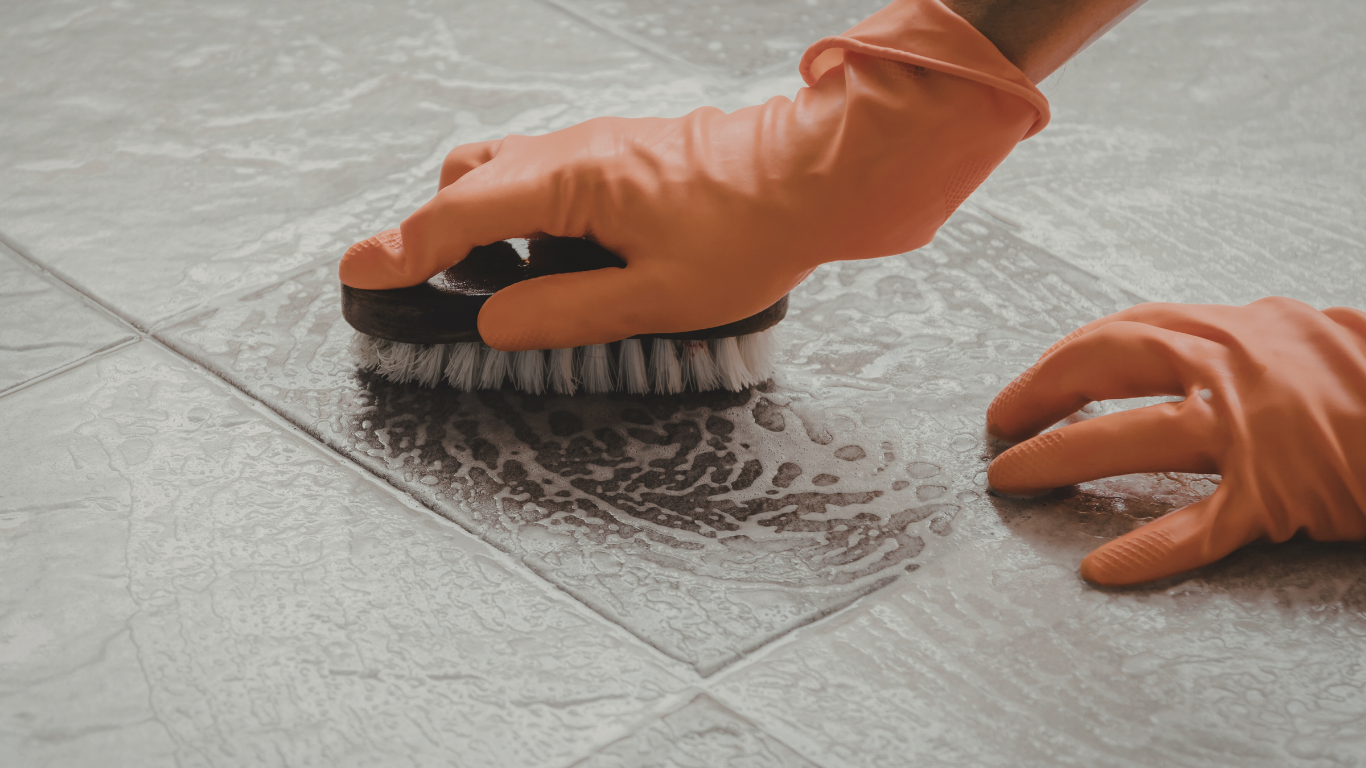 Gloved hands scrubbing floor tiles with a brush; cleaning with suds on a gray surface.