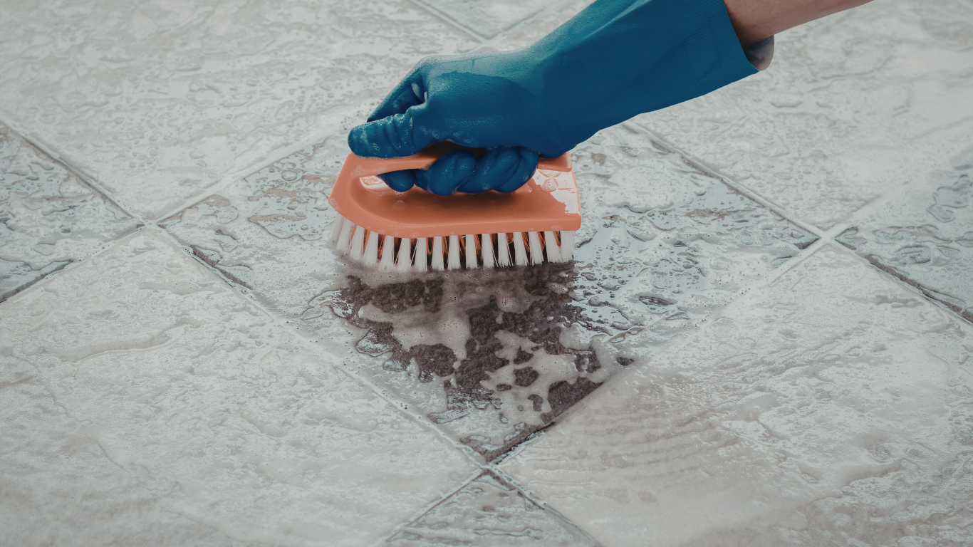 Person wearing a blue glove scrubs a floor tile with a brush, creating soapy foam.