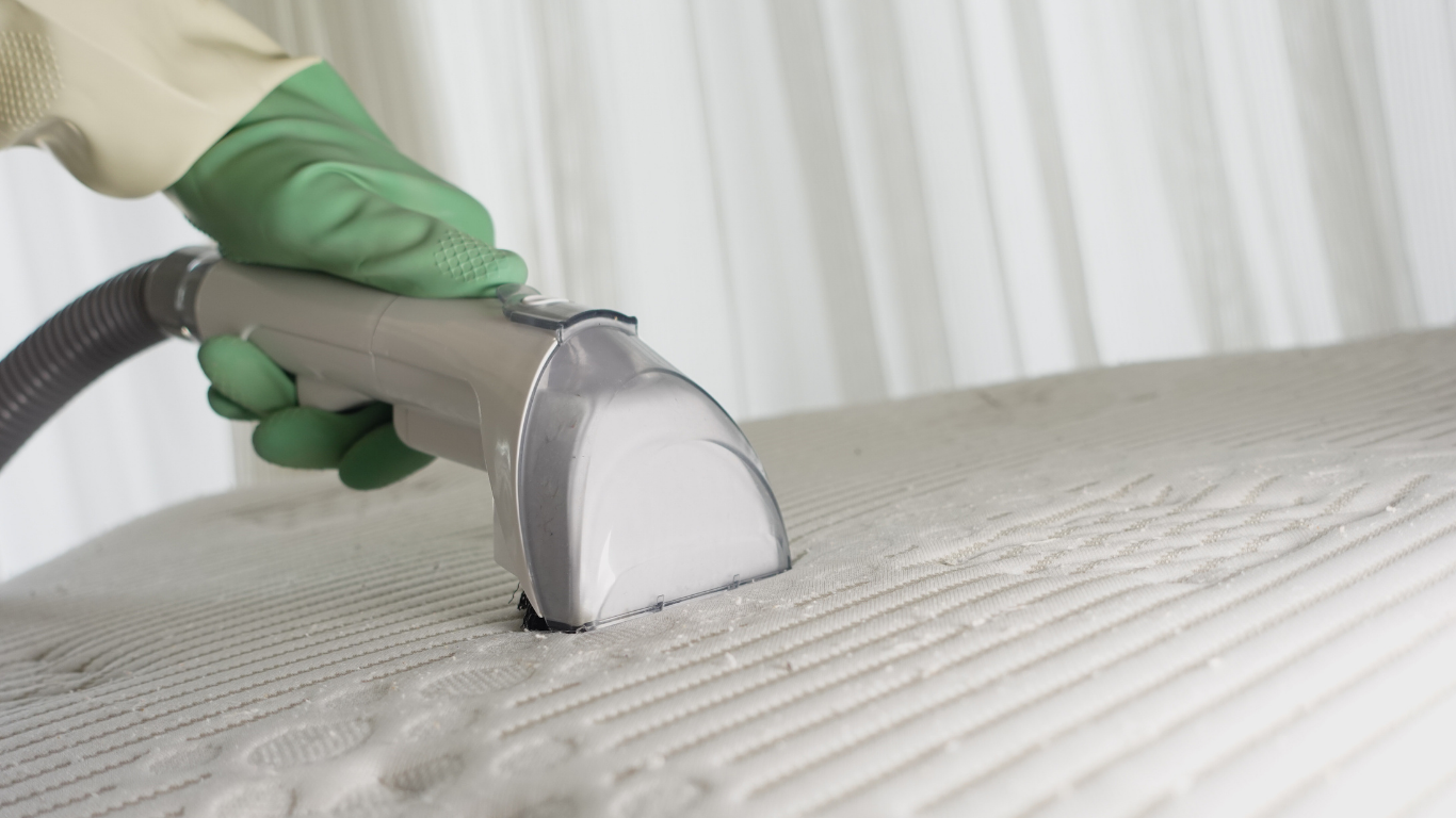 Person wearing green gloves cleaning a white mattress with a handheld vacuum.