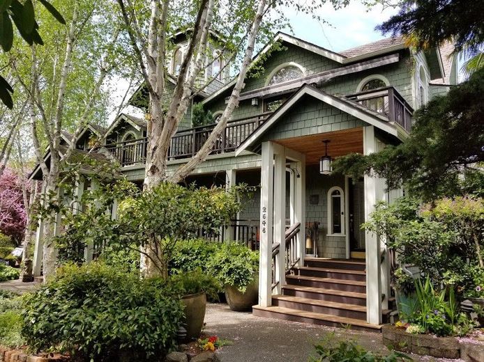 A large green house with a porch and stairs is surrounded by trees and bushes.