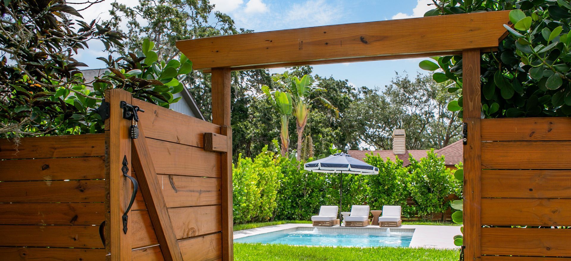 A wooden gate frame opens to a backyard pool area with lounge chairs and a patio umbrella surrounded by greenery.