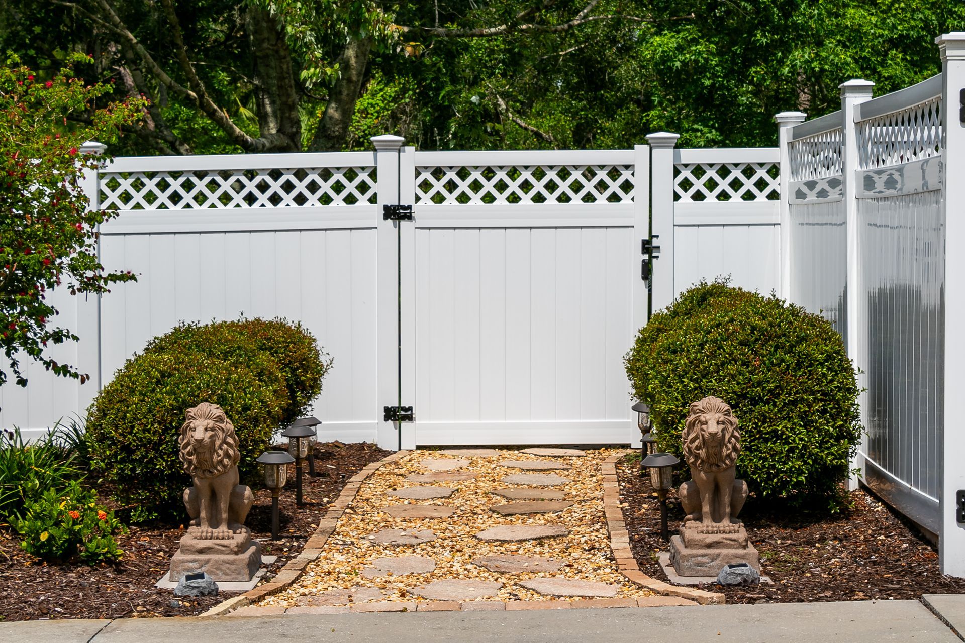 A white vinyl fence with a gate, a stone pathway, two lion statues, and green bushes.