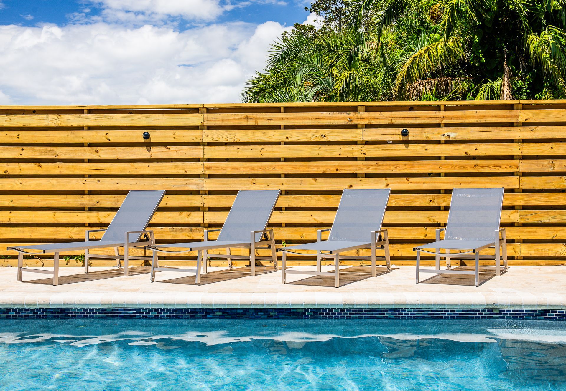 Four gray lounge chairs positioned beside a swimming pool in front of a horizontal wooden fence.