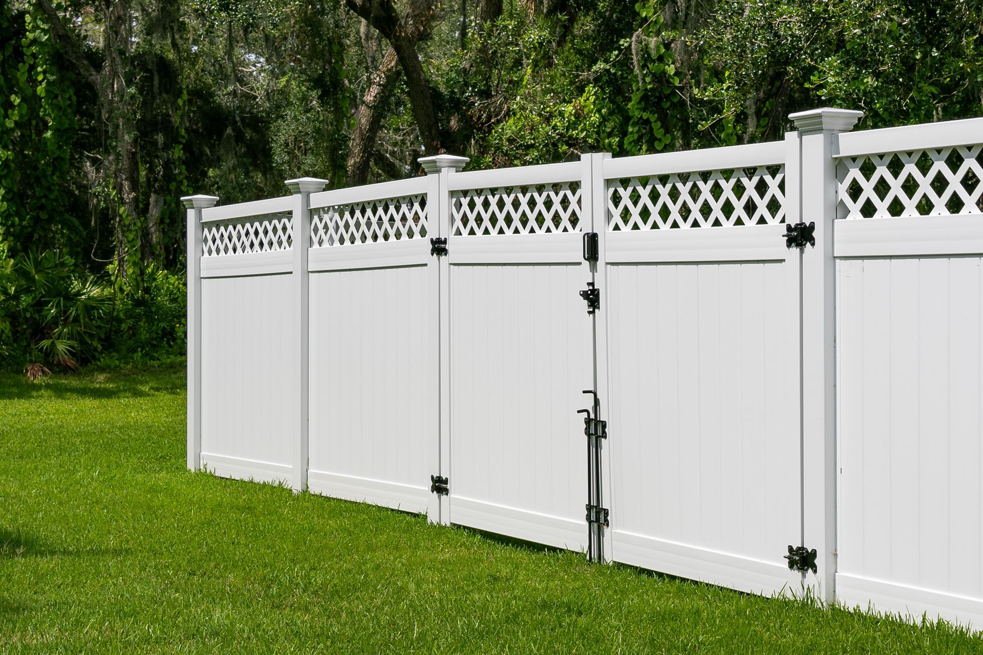 White vinyl fence with a lattice top border and a closed gate, situated on a green lawn in front of trees.