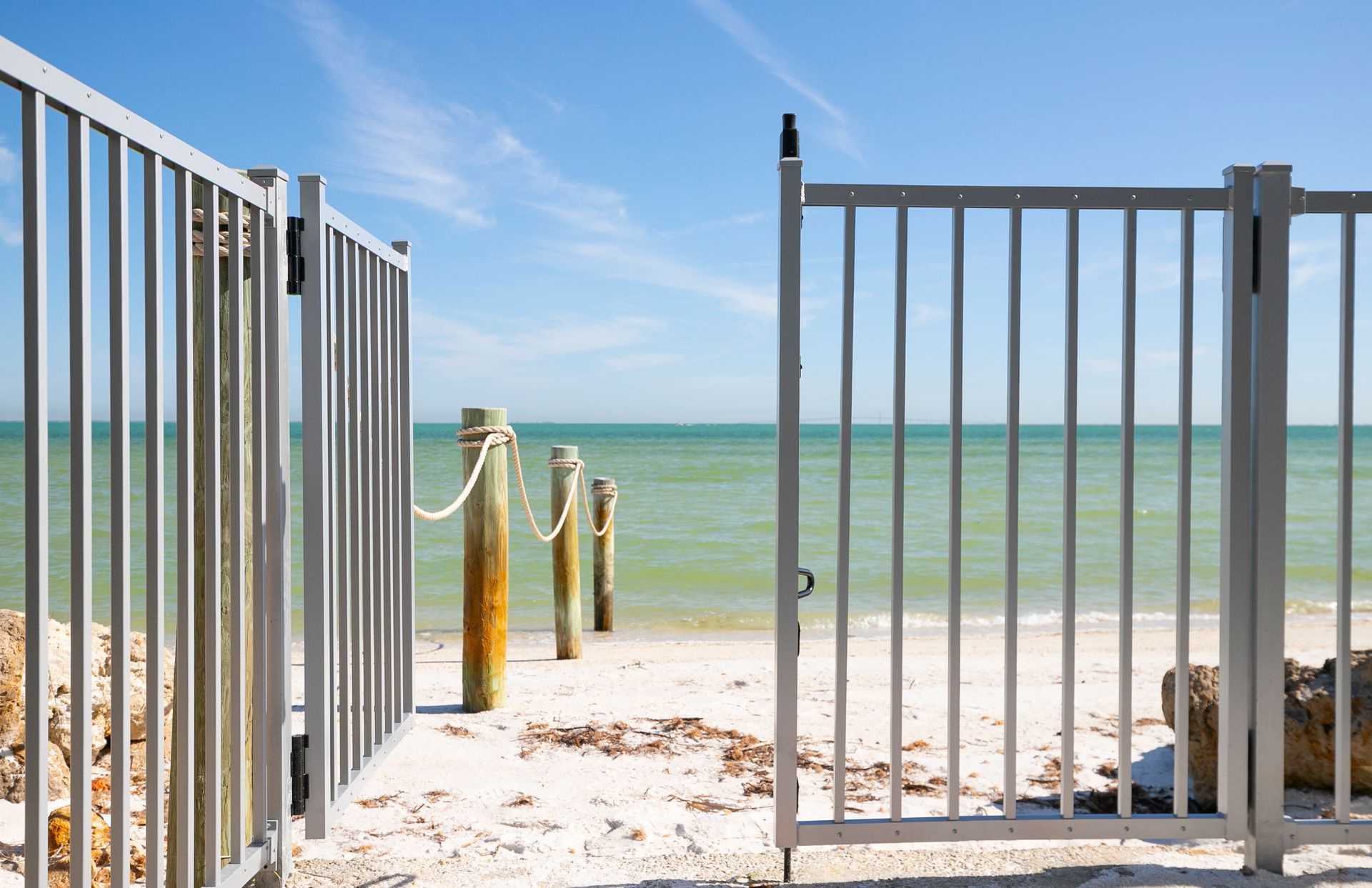An open gate leads to a sandy beach with wooden posts and a rope fence overlooking a calm blue ocean.