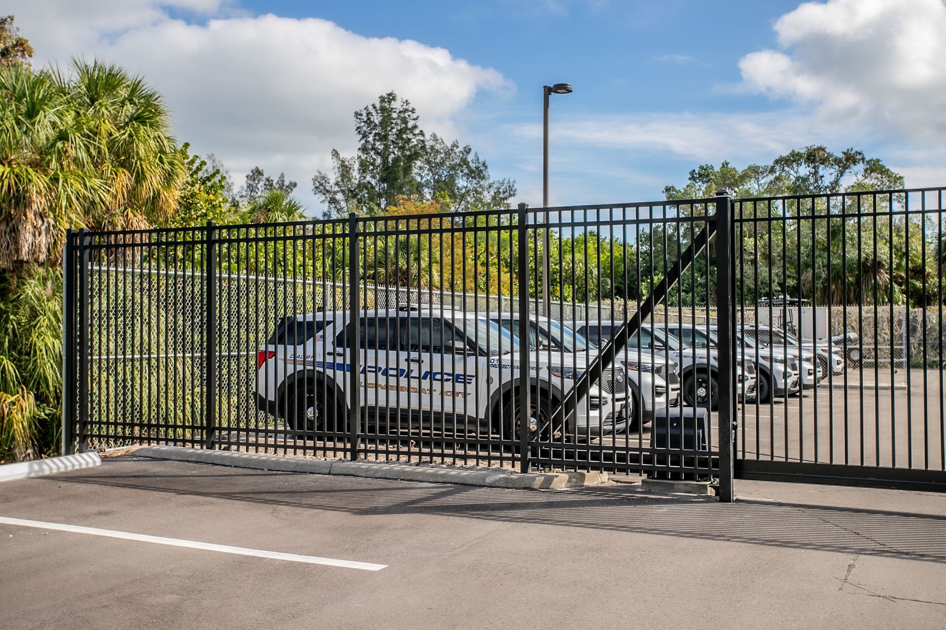 A black metal fence encloses a parking area containing several white police vehicles under a partly cloudy sky.